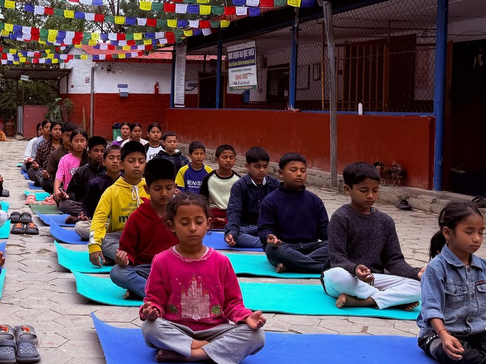 Children at the home sitting cross-legged on mats in the courtyard for morning meditation, with rows of prayer flags overhead.